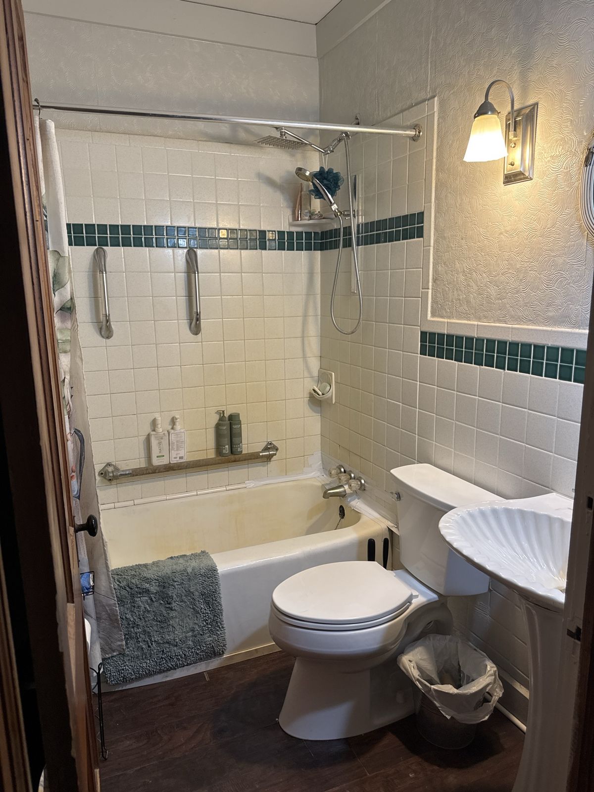 Victorian bathroom before remodeling with dated cream and green accent tile, stained tub, grab bars, and shell pedestal sink