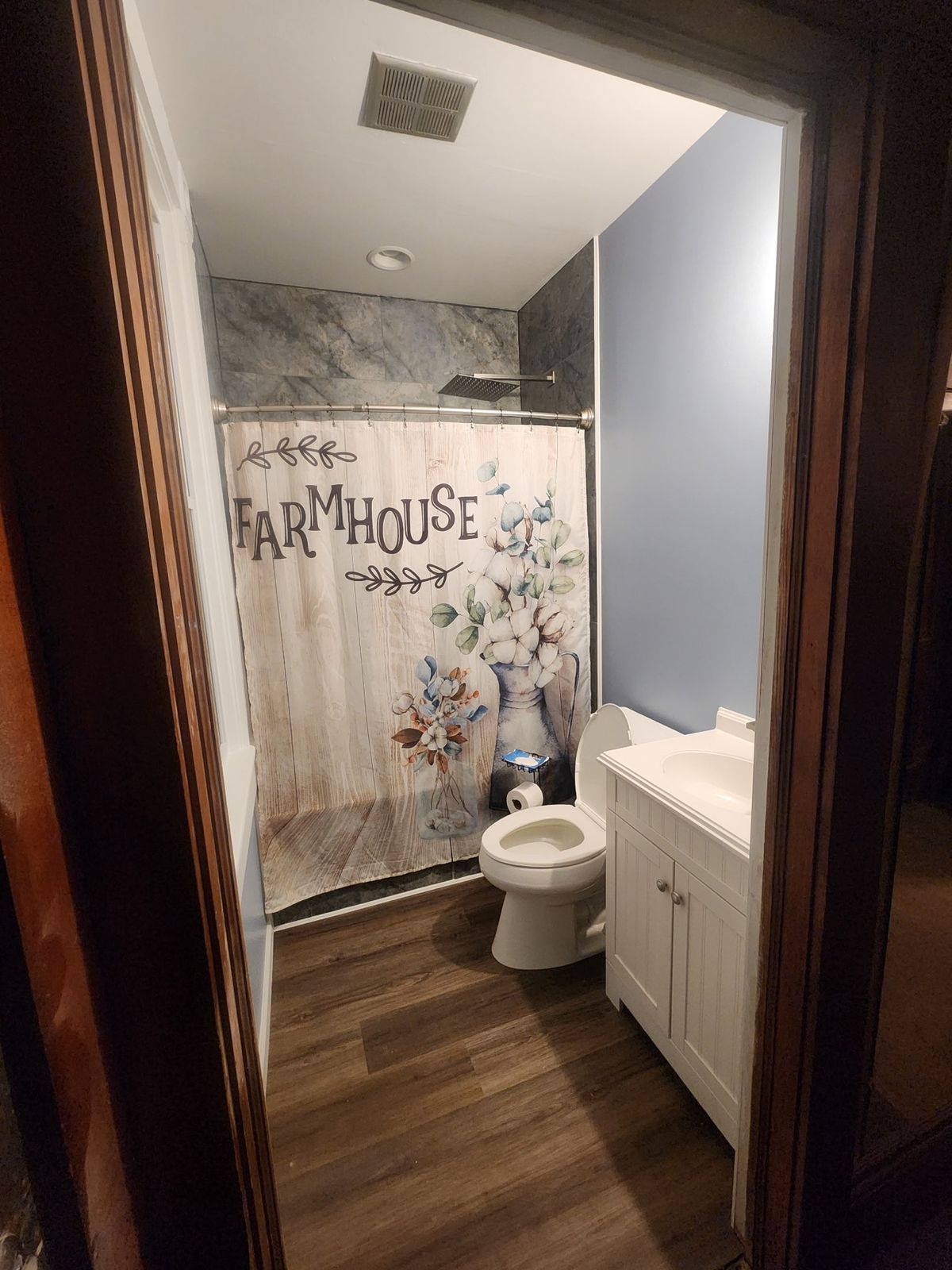 Finished bathroom remodel with farmhouse shower curtain, white vanity, light blue walls, and wood-look flooring