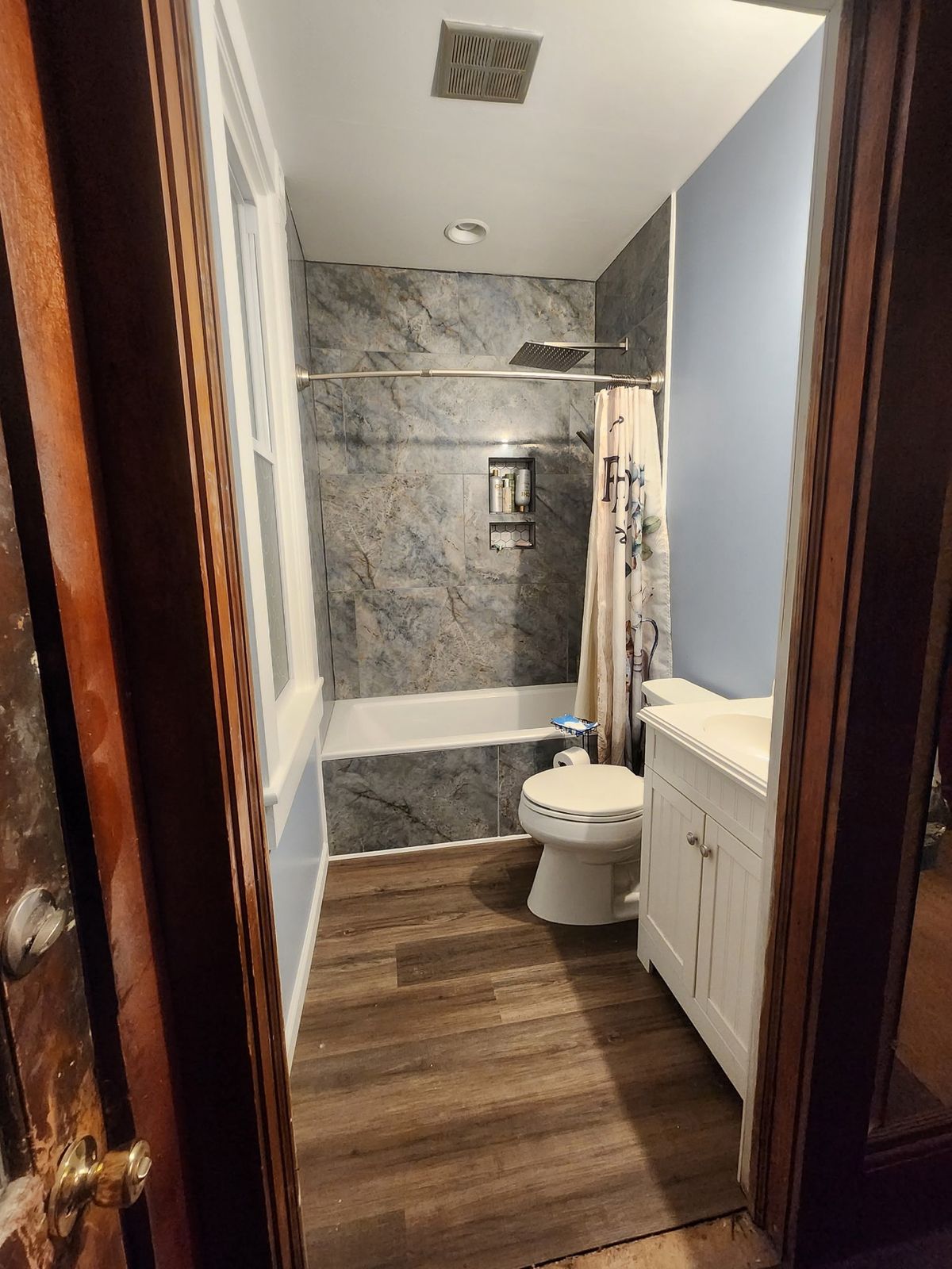 Remodeled Victorian bathroom with marble-look tile tub surround, built-in shower niche, rain shower head, new white vanity, and wood-look flooring